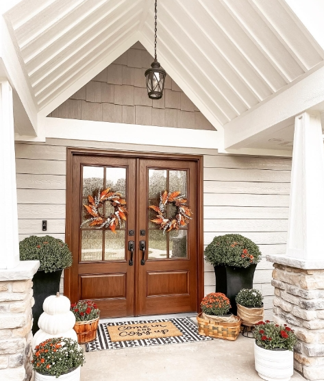 A welcoming porch featuring double wooden doors adorned with vibrant autumn wreaths, surrounded by decorative plants and a cozy doormat that invites guests in.