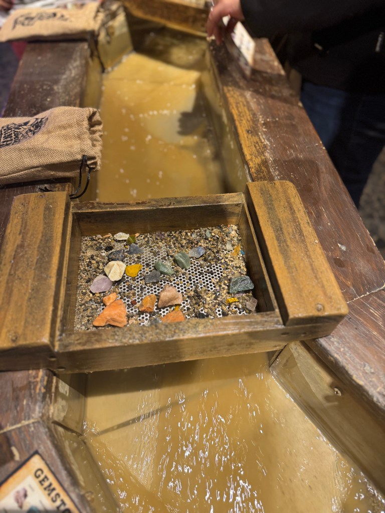 Close-up view of a gemstone mining station with a wooden sluice box filled with gravel and shiny gemstones, set against a backdrop of flowing water.