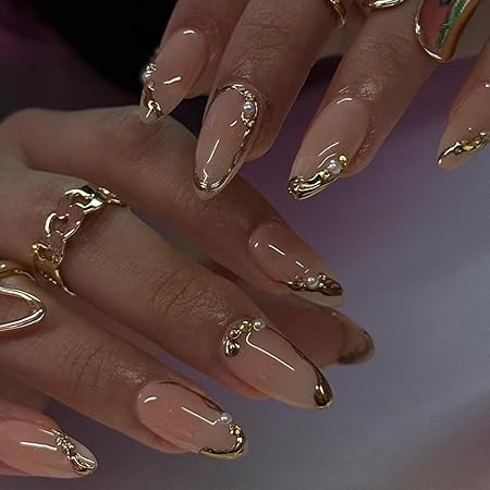 Close-up of elegantly manicured nails featuring a nude polish with gold accents and decorative embellishments, highlighting a sophisticated and glamorous design.