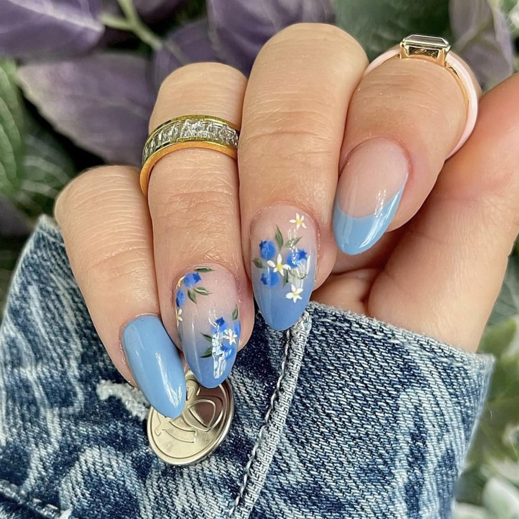 Close-up of a hand showing manicured nails with a light blue design and floral patterns, featuring rings on the fingers and a denim jacket in the background.