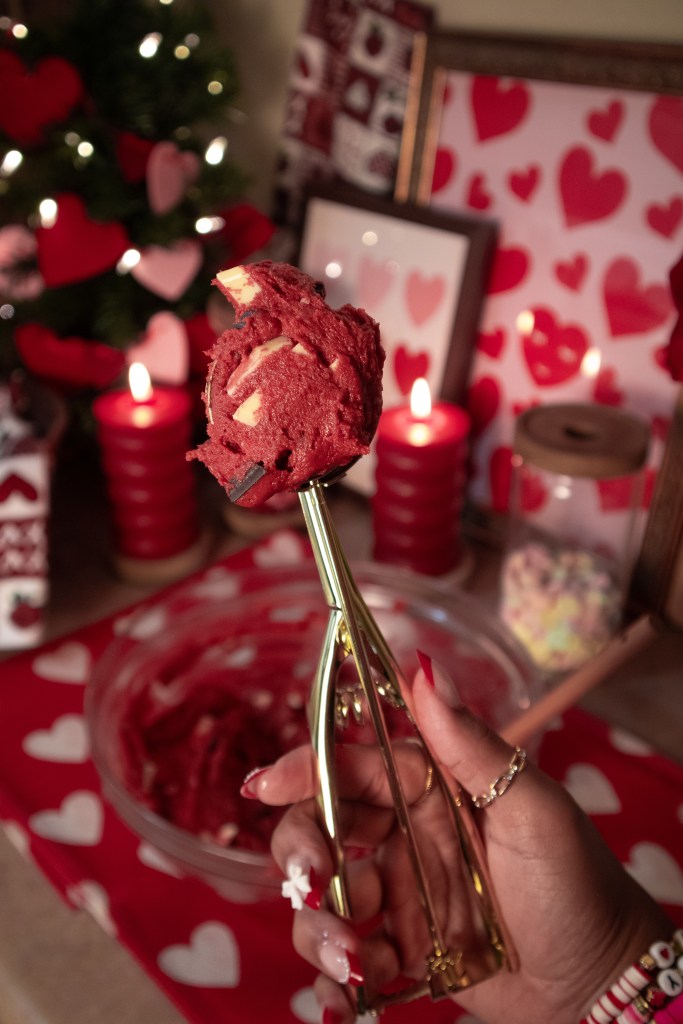 A hand holding a gold ice cream scoop with red cookie dough, surrounded by decorative candles and heart-themed decorations.