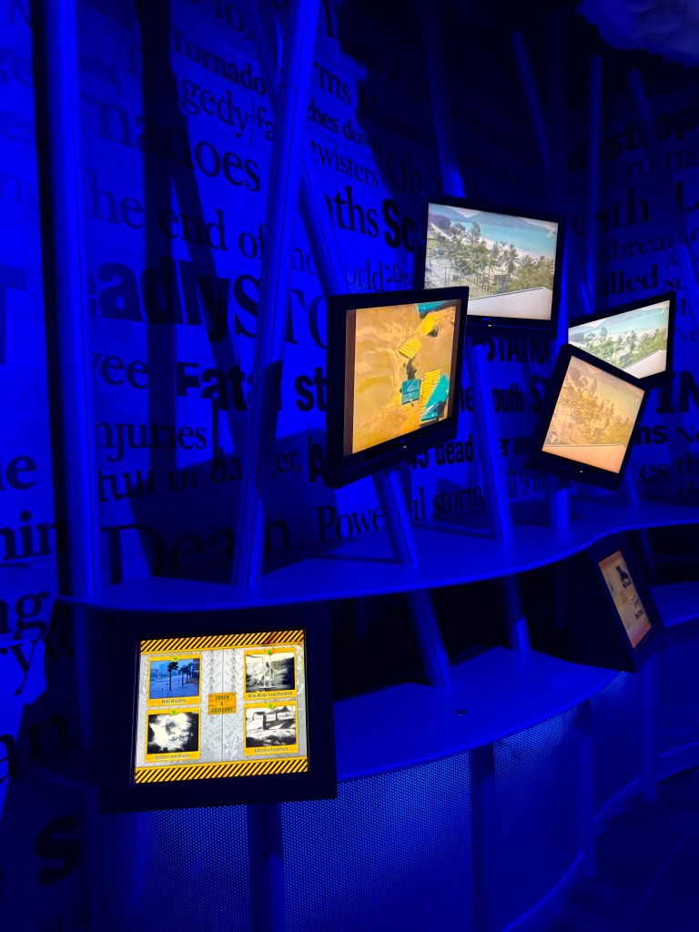 A dimly lit exhibit featuring multiple screens displaying images, surrounded by blue walls with text about severe weather events.