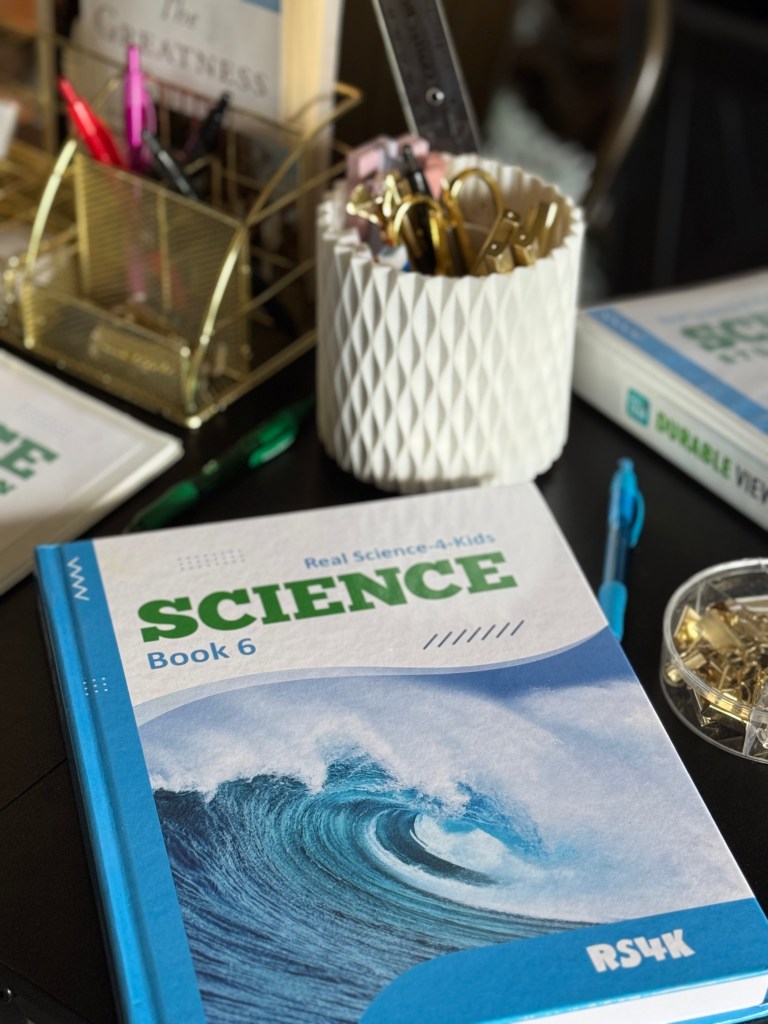 Close-up of 'Real Science 4 Kids Book 6' on a table, showcasing its cover featuring a wave design, surrounded by pens, paperclips, and notebooks.