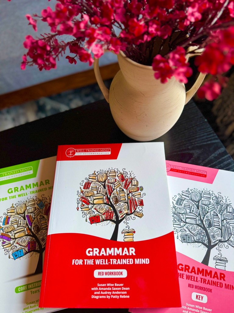 Three grammar workbooks from 'Grammar for the Well-Trained Mind' are displayed on a table. The main workbook is red, featuring illustrations of books arranged in a tree shape. A vase with pink flowers is also in the scene.