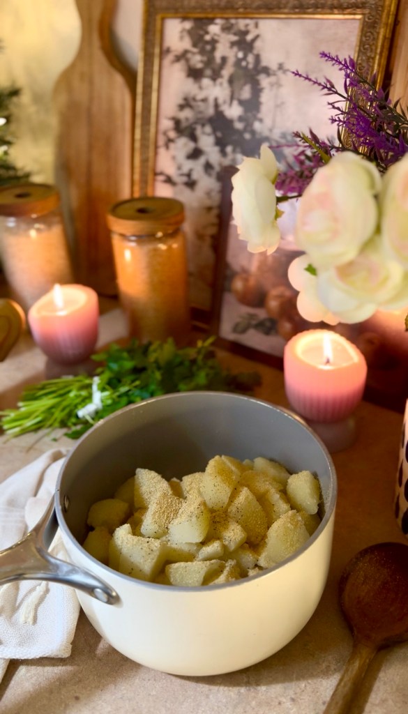 A pot filled with chopped potatoes seasoned with spices, surrounded by candles, decorative jars, flowers, and a wooden spoon on a countertop.