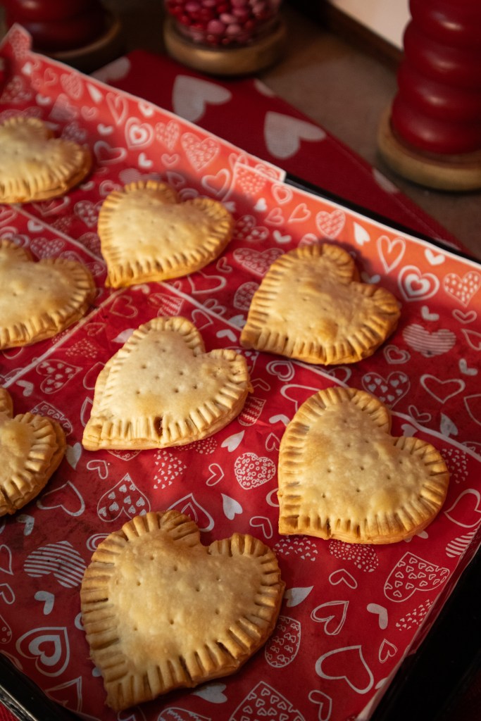 A tray of heart-shaped pastries placed on red paper with heart patterns, featuring a golden-brown crust.