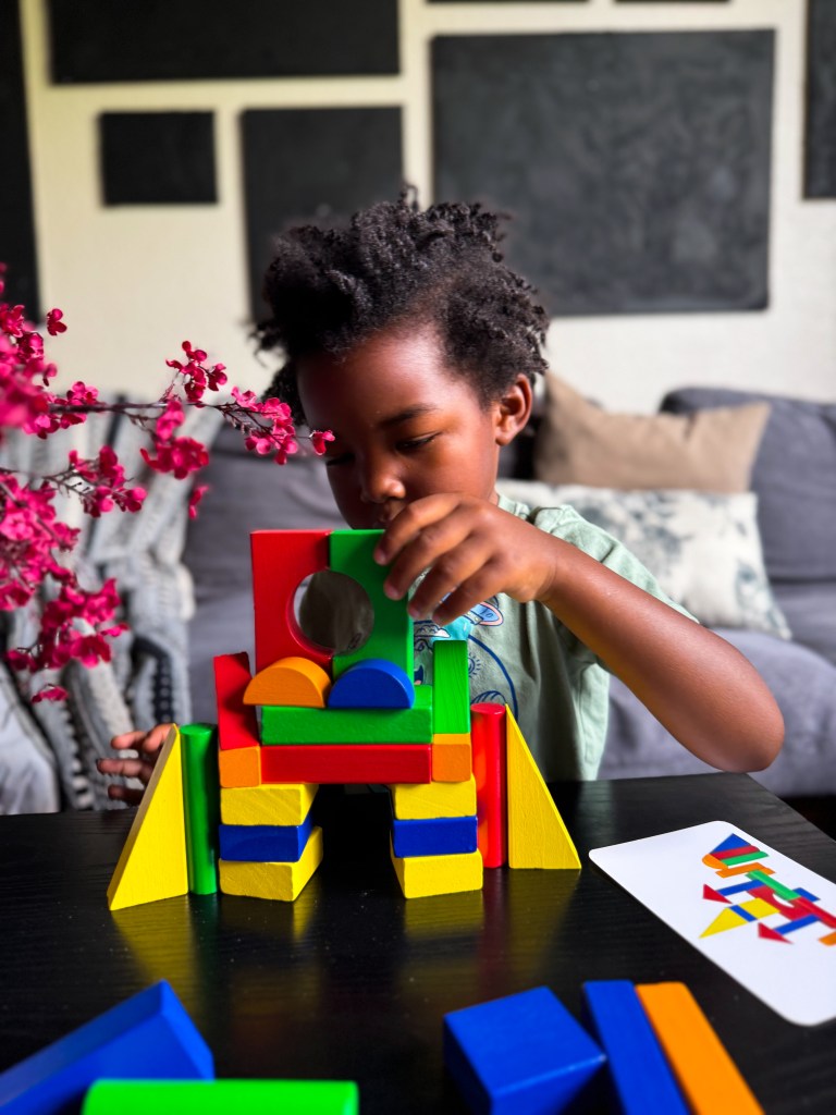 A young child playing with colorful building blocks, constructing a structure. The child is focused on placing a green block on the red base, surrounded by various shaped blocks and a card with colorful shapes.