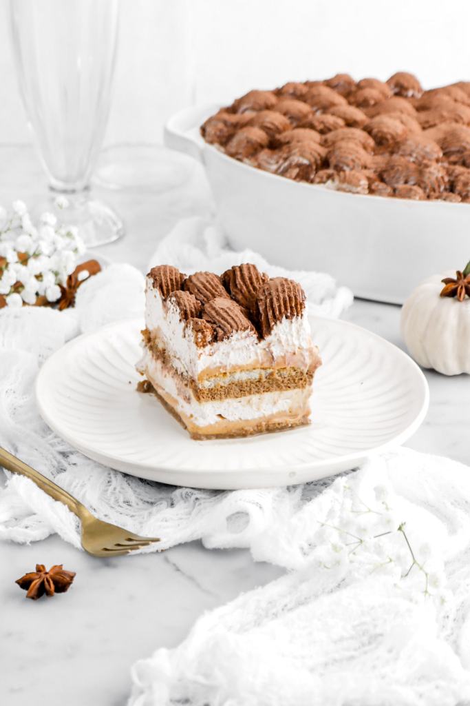 A slice of creamy layered pumpkin dessert with whipped topping on a white plate, surrounded by decorative fall elements like a small pumpkin and star anise.