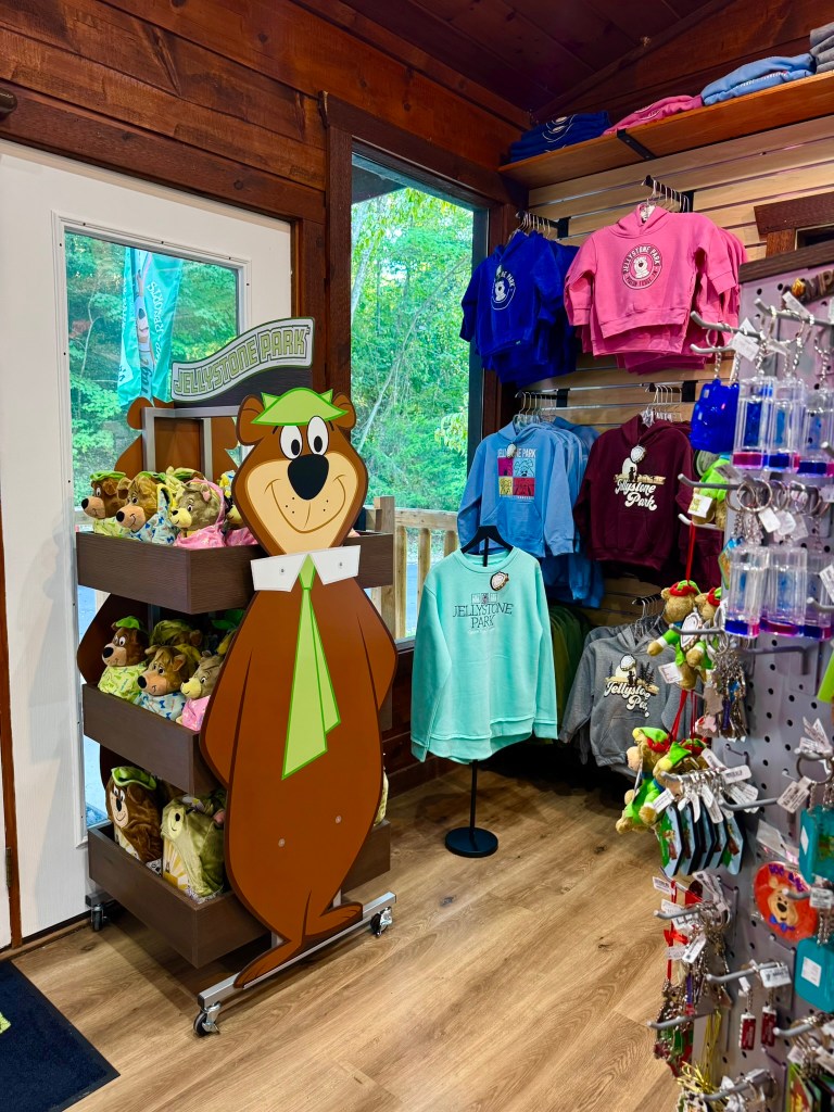 Merchandise display featuring a cartoon bear character at a Jellystone Park store, with clothing and plush toys visible inside a wooden interior.