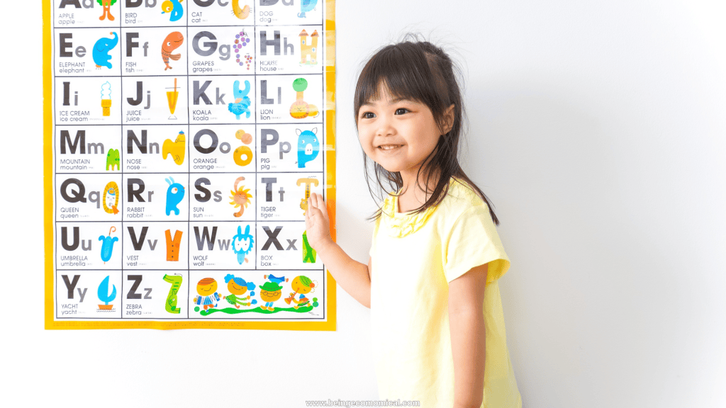 Preschool-aged child standing beside an alphabet chart while practicing letter recognition.
