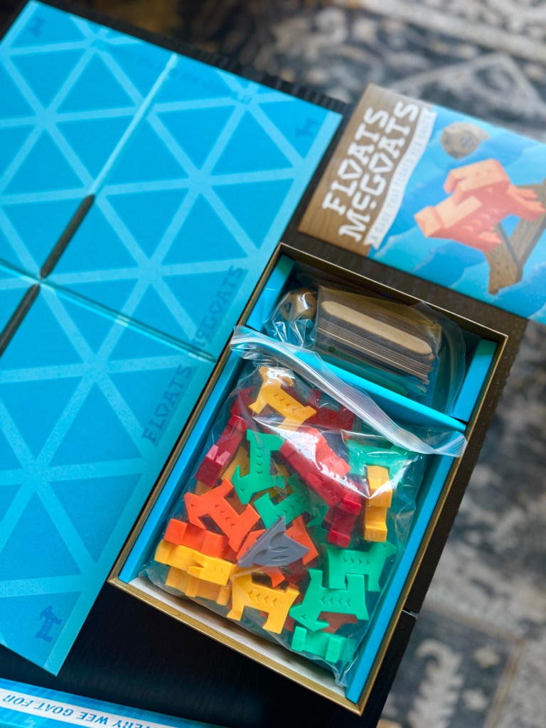 Open game box of 'Floats McGoats' featuring colorful goat-shaped pieces, a wooden game piece, and a rule booklet on a blue background.