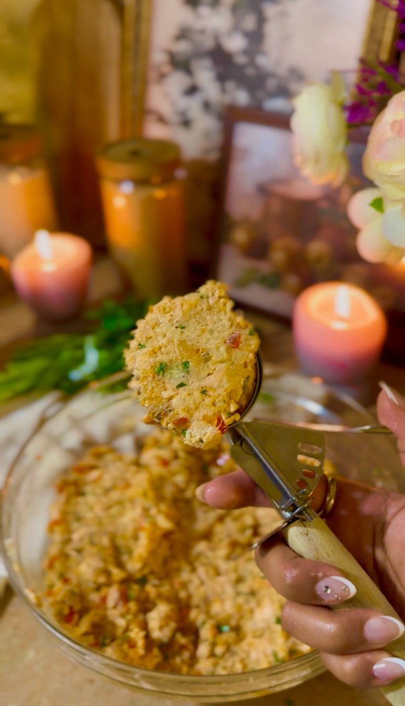 A hand holding a cookie scoop filled with a mixture, with a bowl of the same mixture in the background and candles and framed pictures adding ambiance.