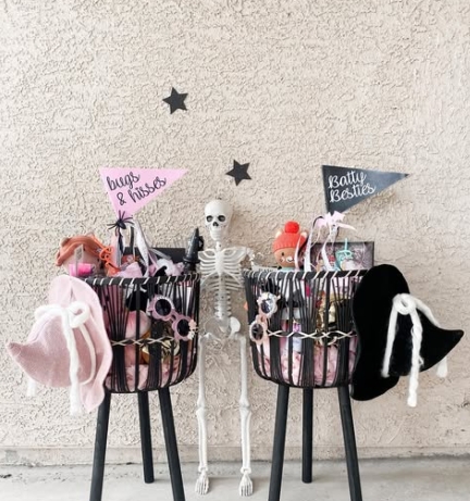 Two black wire baskets filled with Halloween goodies and themed accessories, accompanied by a skeleton and small decorative pennants reading 'Bugs & Kisses' and 'Batty Besties'.