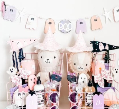Two pastel pink wire baskets filled with Halloween-themed goodies, including plush ghost dolls wearing party hats, decorated bottles, and playful pennants, surrounded by a festive 'BOO' banner.