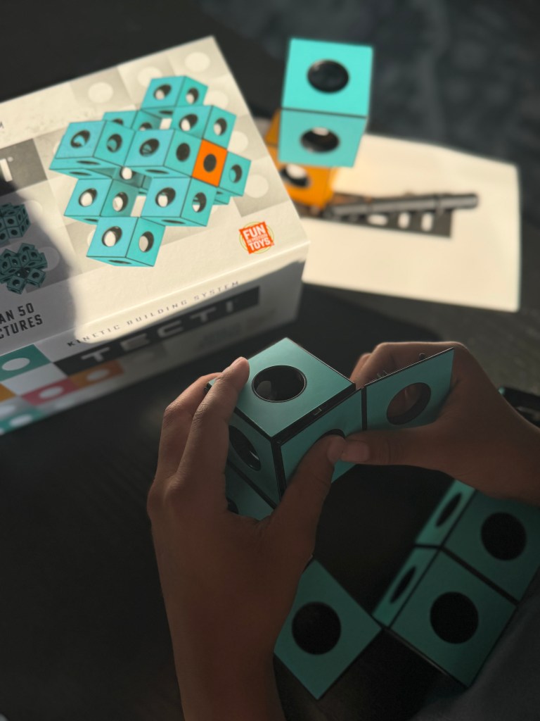 A child assembling a turquoise Tecti kinetic building block set, with the product box and instruction booklet in the background.