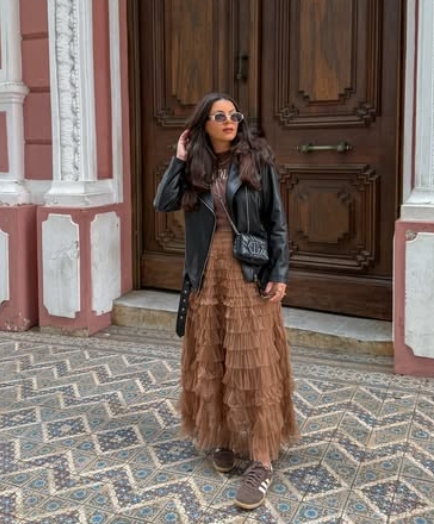 A woman with long hair stands outside a door, wearing a black leather jacket over a tiered brown maxi dress and brown sneakers. She accessorizes with round sunglasses and holds a small bag.