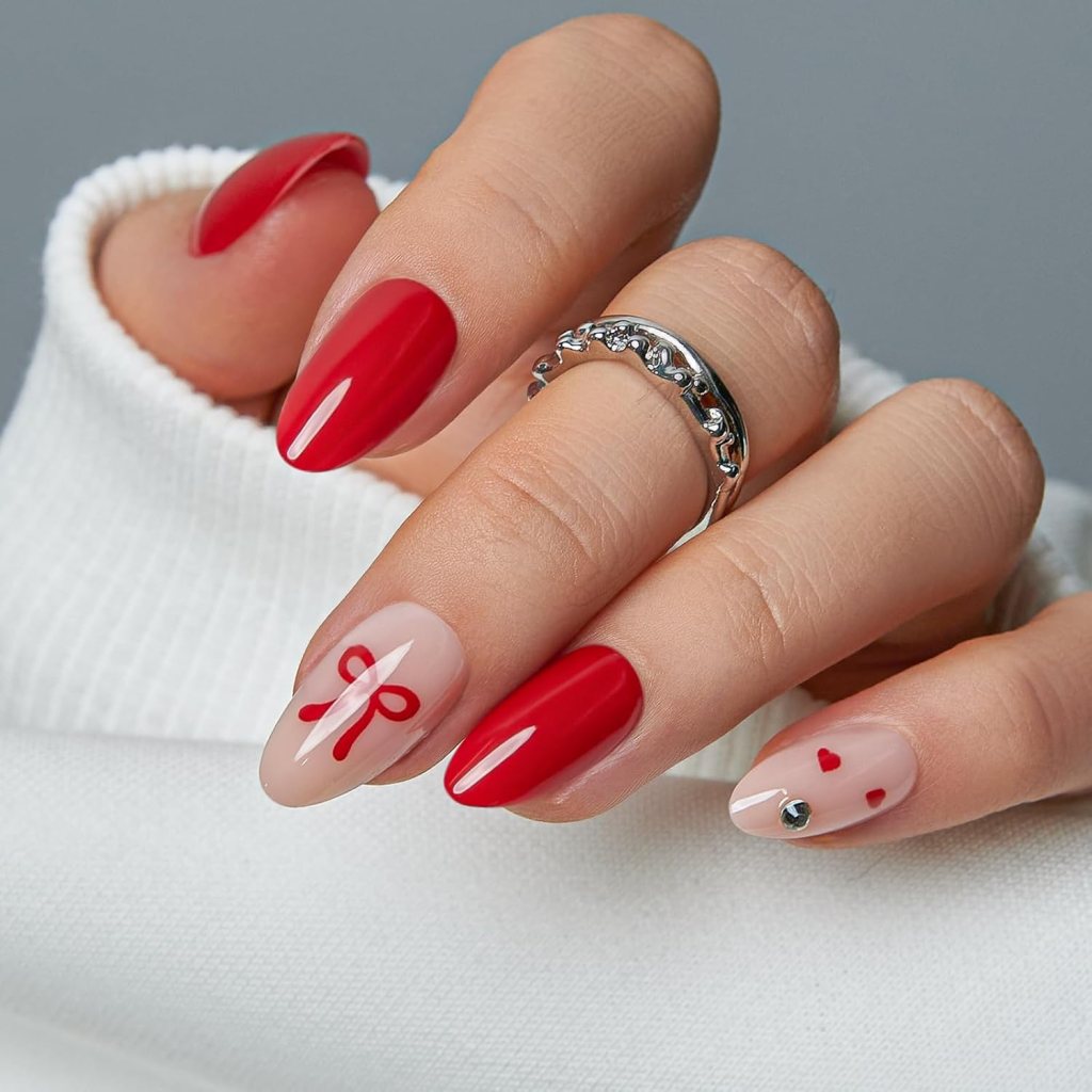 A close-up of a hand with stylish nails, featuring a mix of bold red and nude polish, decorated with red bows and hearts, along with a silver ring on the ring finger.