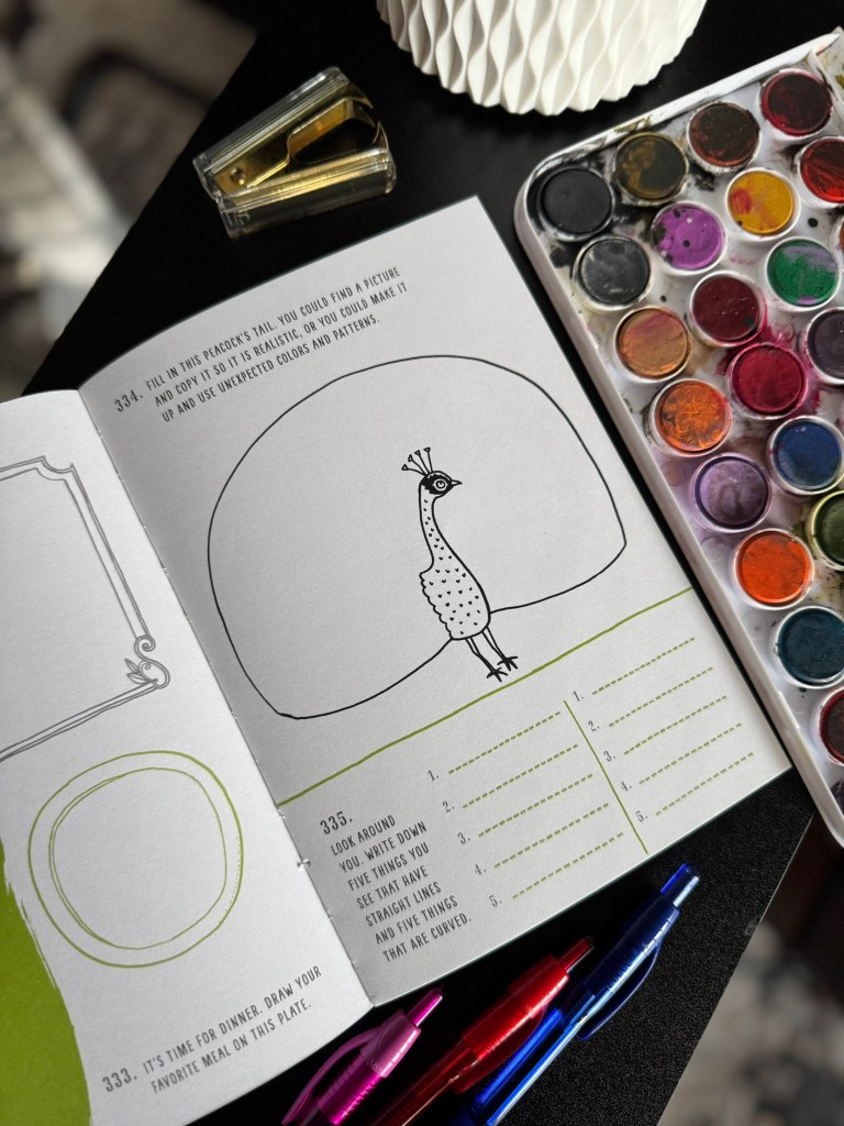 An open art prompt book page featuring a drawing of a peacock with instructions for creative exploration, alongside watercolors, pens, and a stapler on a black table.