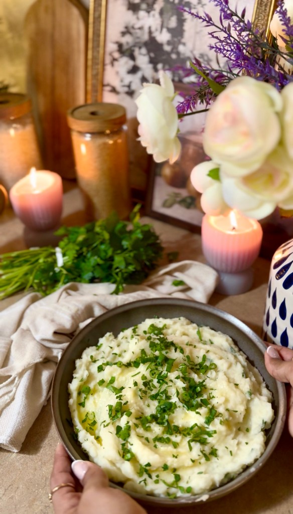A person holding a bowl of creamy mashed potatoes topped with chopped green herbs, surrounded by candles, flowers, and decorative items.