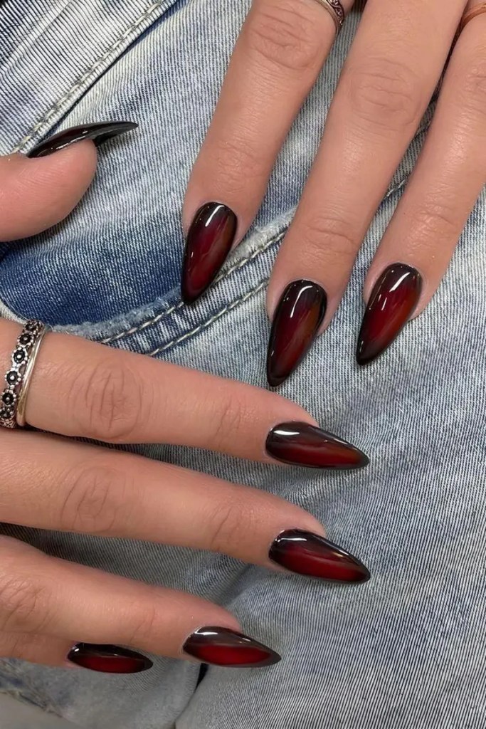 Close-up of hands displaying long, pointed nails painted in a deep red and black gradient, resting on a denim fabric.