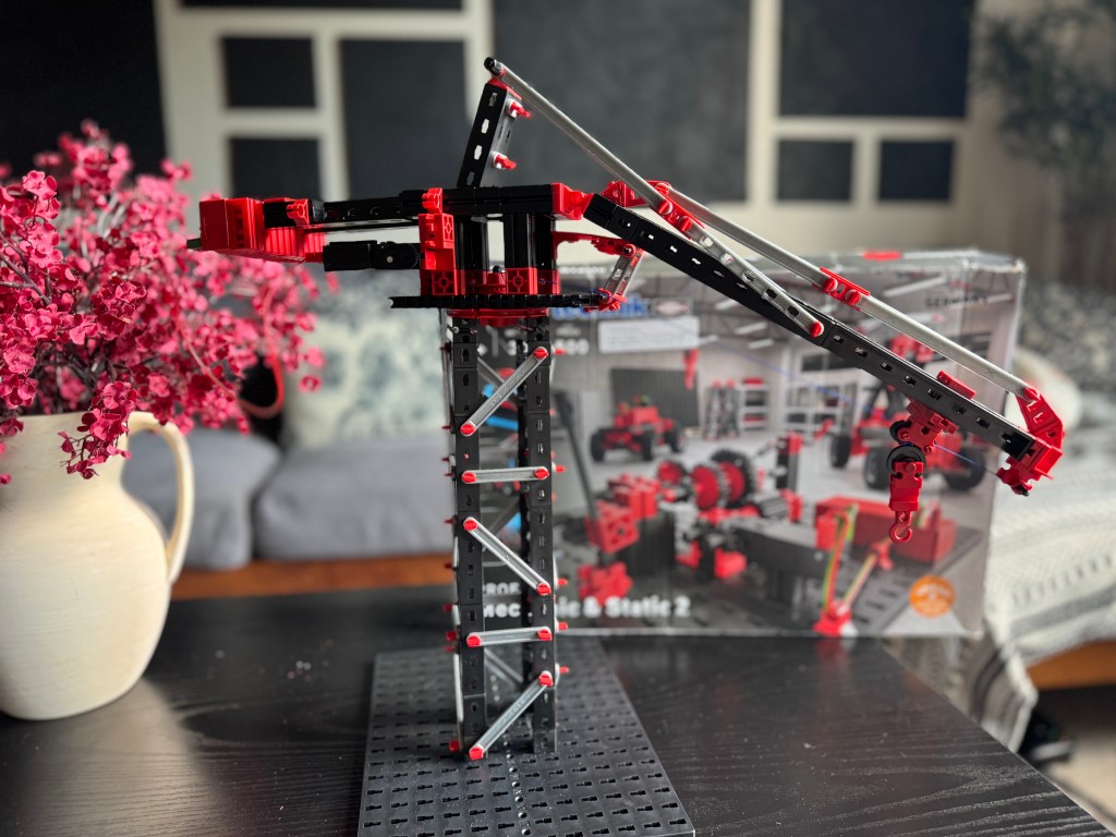 A Fischer Technik STEM kit crane model built with colorful pieces, placed on a black building board beside a pink flower arrangement, with a background showing parts of the kit's box.