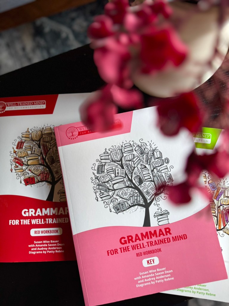 Close-up of two educational workbooks titled 'Grammar for the Well-Trained Mind,' featuring illustrations of books and a tree motif, with a floral arrangement in the foreground.