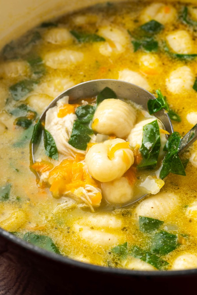 A ladle scooping creamy chicken soup with gnocchi and spinach from a pot, showcasing a warm and comforting dish.