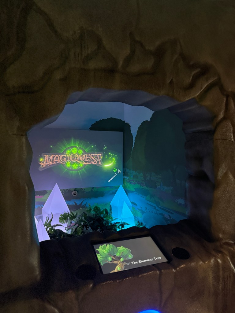 A view through a stone archway of the MagiQuest interactive adventure area at Great Wolf Lodge. The wall features glowing graphics of the MagiQuest logo, along with decorative elements like blue crystal-like structures and lush greenery.