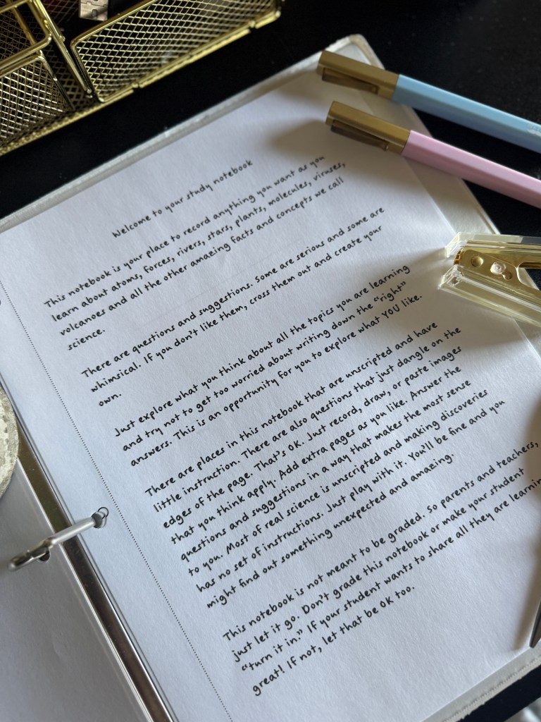 A close-up of an open study notebook with text on a page explaining its purpose, surrounded by colorful pens and a metal pen holder.