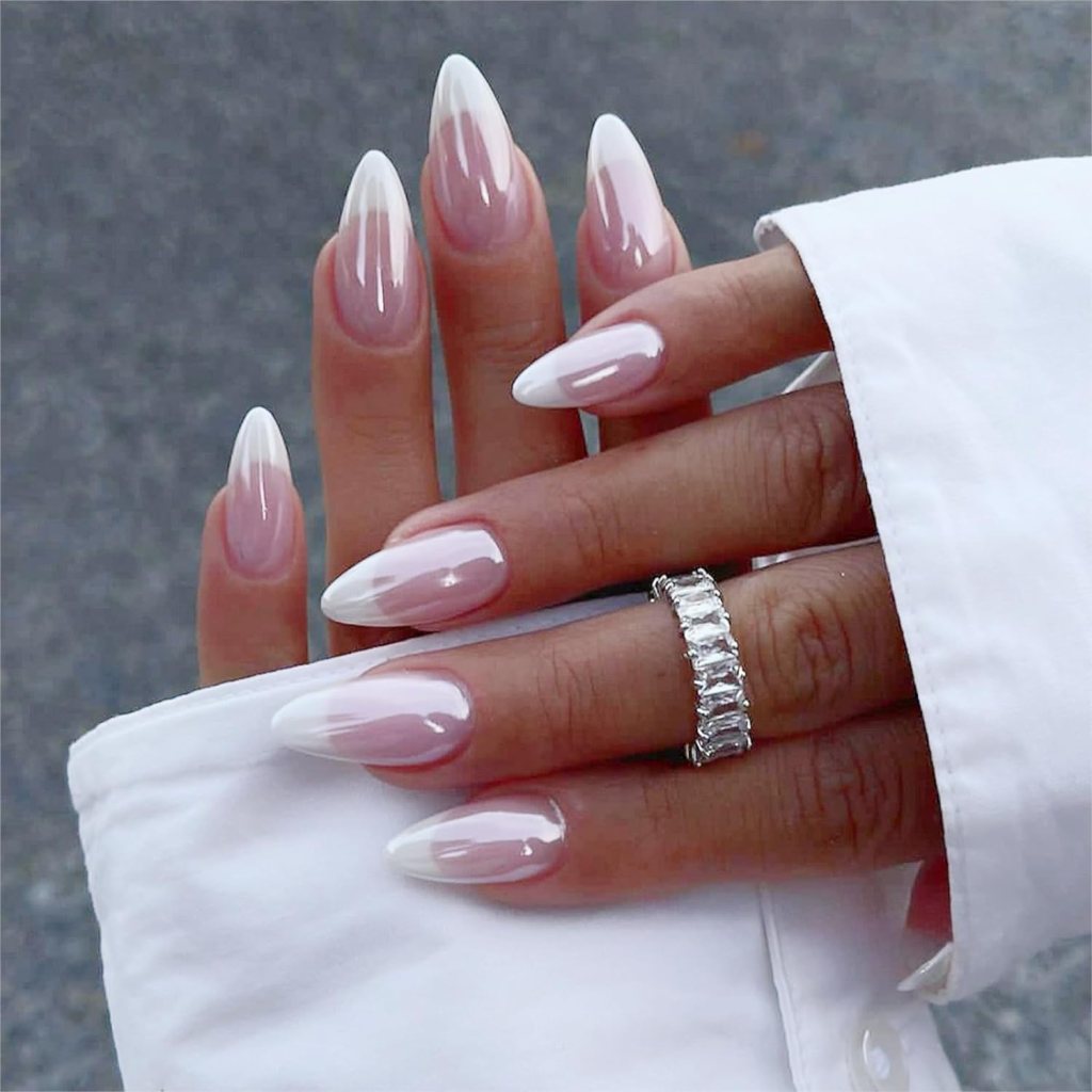 Close-up of elegant, long almond-shaped nails with a gradient ombre effect, featuring a soft pink base with white tips. A silver ring is worn on one finger, and the hand rests against a white shirt.