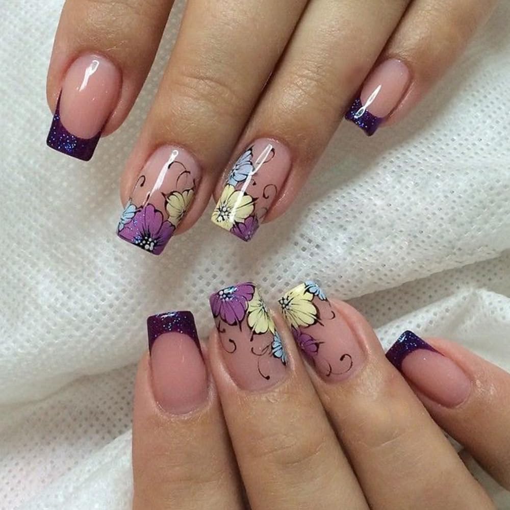 A close-up view of elegantly manicured nails featuring a natural pink base with colorful floral designs in purple, yellow, and blue, complemented by dark purple glitter tips.