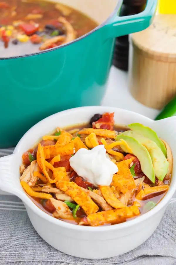 A white bowl filled with Chicken Tortilla Soup, topped with shredded cheese, tortilla strips, a dollop of sour cream, and avocado slices, with a green Dutch oven in the background.