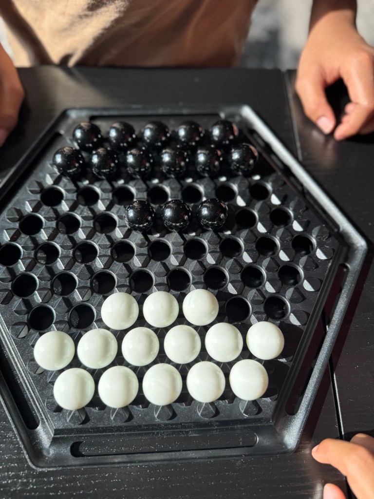 A close-up view of an Abalone game board featuring black and white marbles arranged in a hexagonal grid, with hands of players visible at the edges.