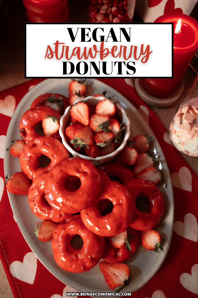 A plate of vegan strawberry donuts glazed with a bright red icing, accompanied by fresh strawberries. The plate is set on a heart-patterned tablecloth, with decorative candles and other colorful accents in the background.