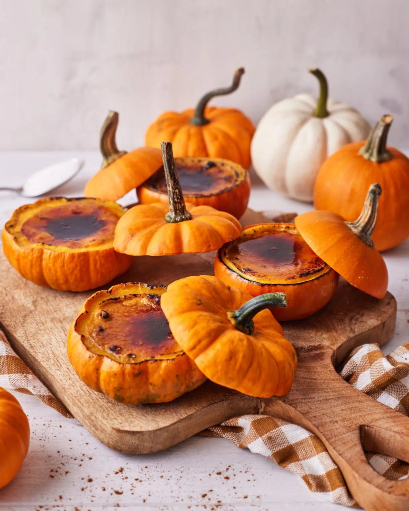Mini pumpkin crème brûlées served in hollowed-out pumpkins on a wooden cutting board, with an orange and white pumpkin in the background.