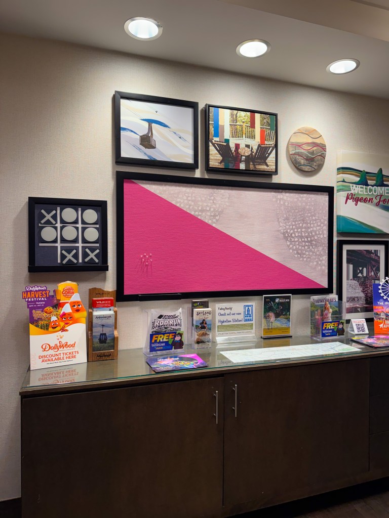 A hotel lobby display featuring a large pink and white artwork, surrounded by several framed pictures, including landscapes and abstract art. A wooden cabinet with multiple brochures and promotional materials for local attractions and events is in the foreground.
