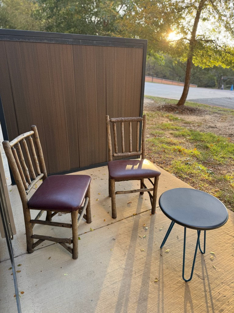 A small outdoor seating area with two rustic wooden chairs and a round black table, surrounded by trees and a hint of sunlight in the background. Great Wolf Lodge in Grapevine, TX. 