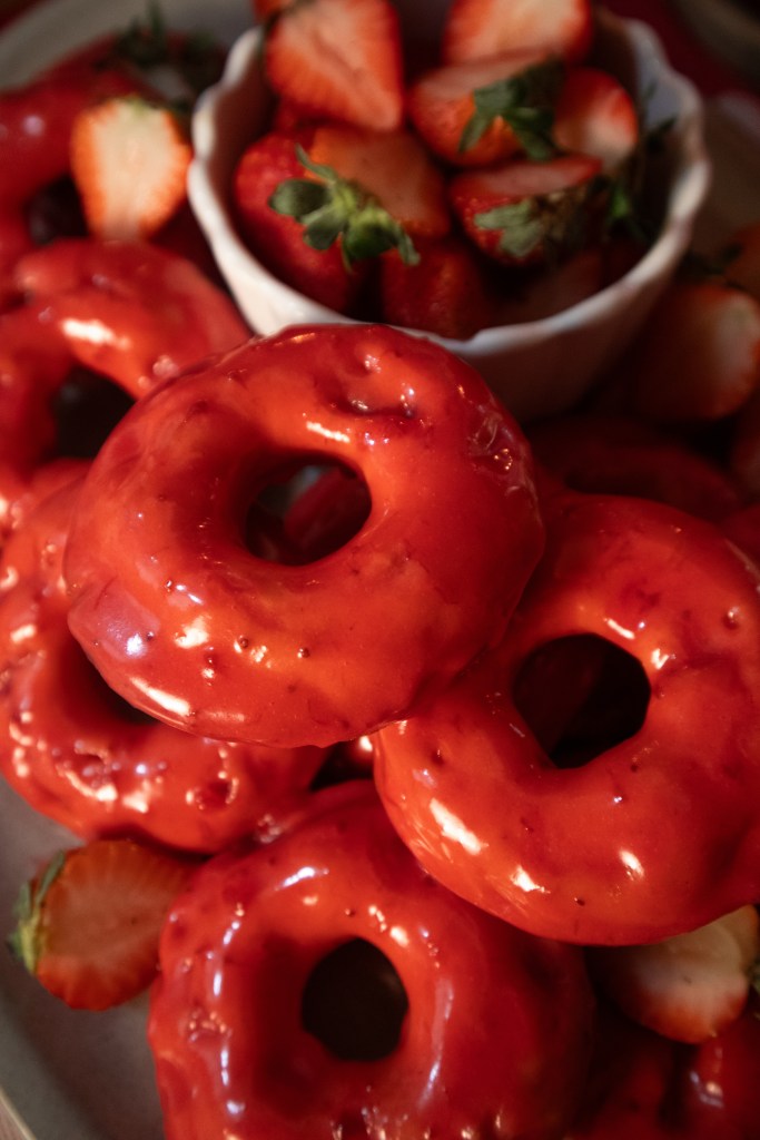 A close-up of glazed red donuts arranged on a plate, with a small bowl of sliced strawberries in the background.