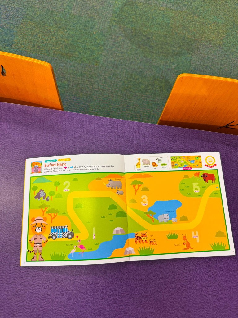 An open children's activity book titled 'Safari Park' showing a colorful illustration of a path with numbers and various animal stickers, including a giraffe, elephants, and lions, on a vibrant green and yellow background.