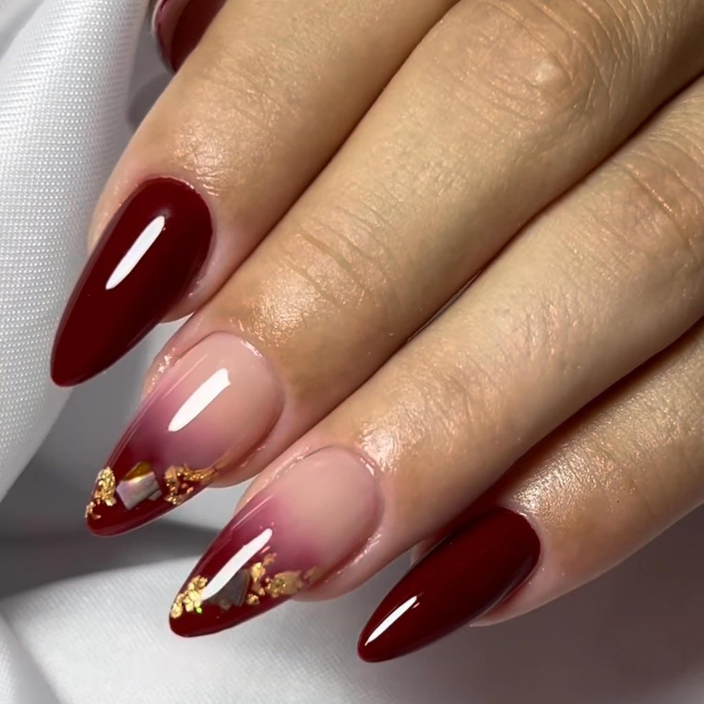 Close-up of elegantly manicured nails with a gradient of burgundy to nude, adorned with gold flakes and a glossy finish.