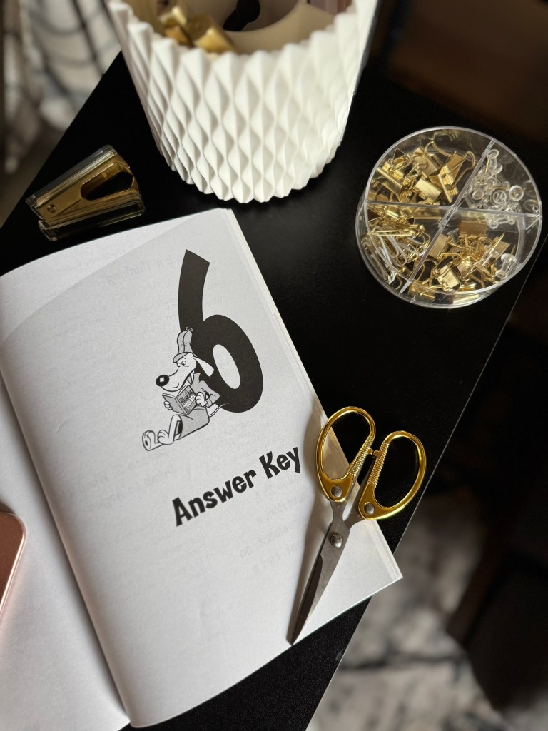 Open book page featuring the title 'Answer Key' with a cartoon dog illustration, a pair of scissors, a stapler, and a container of paper clips on a black table.