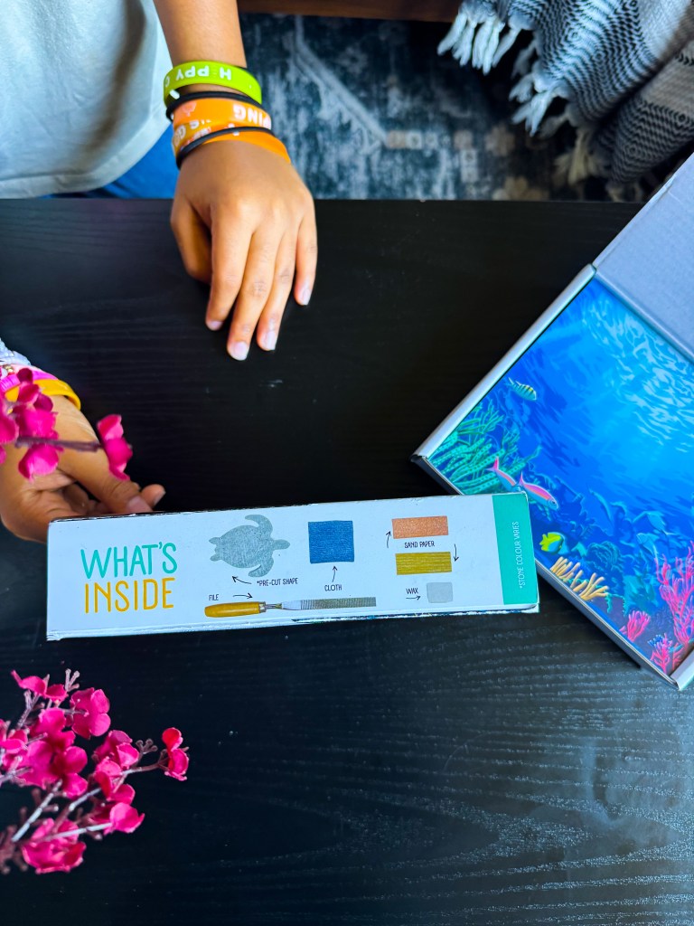 An overhead view of a child's hand holding a box labeled 'What's Inside,' displaying items such as a file, pre-cut shape, cloth, and sandpaper. A colorful marine-themed illustration is visible on the box. A small decorative flower arrangement is in the foreground.