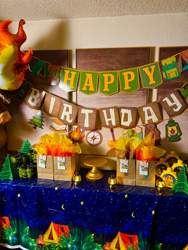 Happy birthday camping party banner displayed above favor bags and lantern decorations.