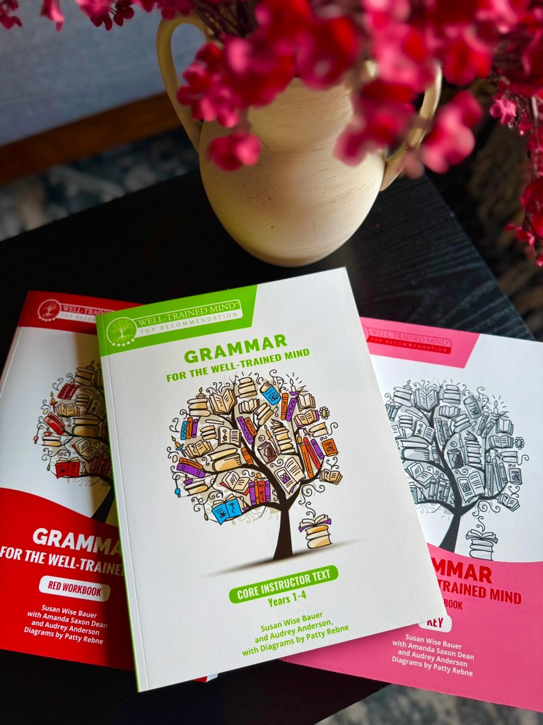 A close-up view of three grammar textbooks from 'Grammar for the Well-Trained Mind', including the Core Instructor Text and two workbooks, displayed beside a decorative vase with pink flowers.