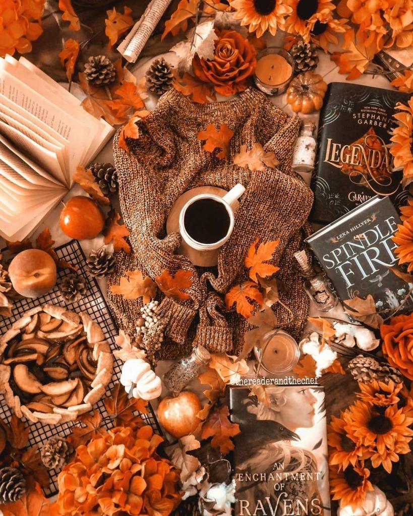 A cozy fall-themed flat lay featuring a knitted sweater, a mug of coffee, books, and seasonal decorations like autumn leaves, sunflowers, pinecones, and pumpkins.