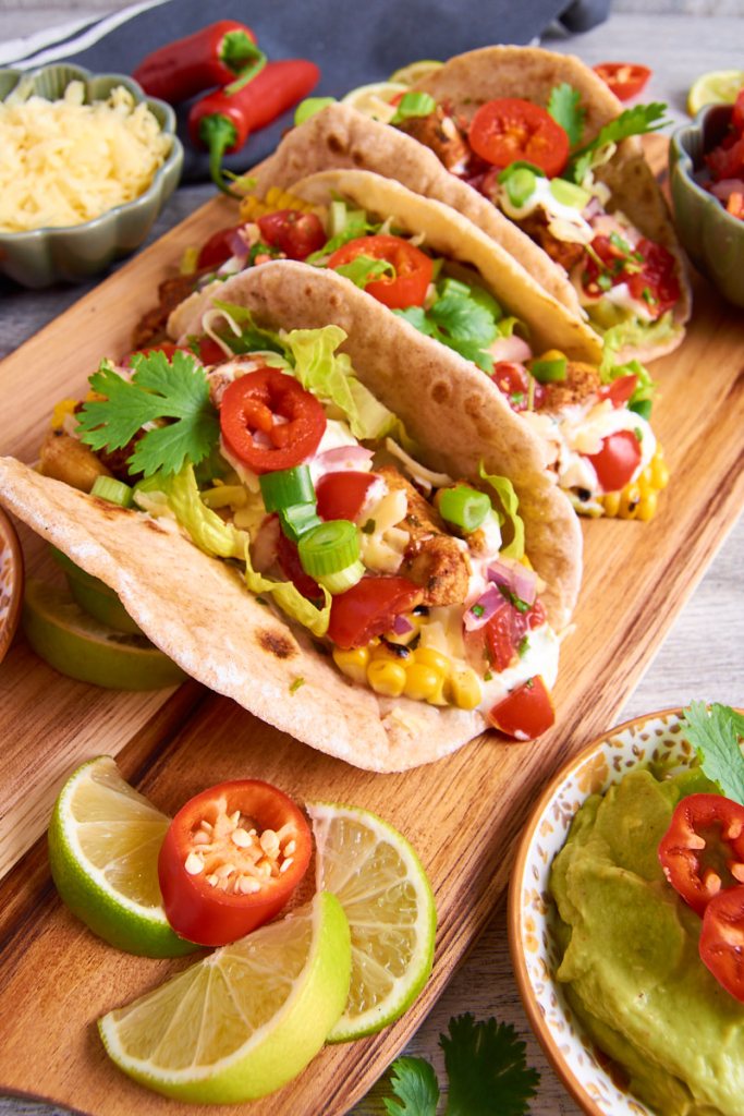 Three chicken tacos filled with toppings like lettuce, tomato, and cilantro, served on a wooden platter with lime wedges and a bowl of guacamole.