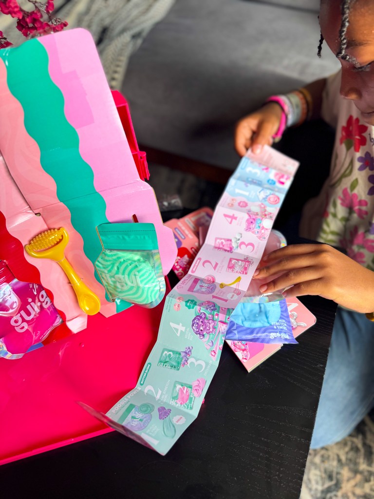 A child is playing with a colorful set that includes containers and a sequence of cards with numbers and illustrations. The set is arranged on a black table with a pink carrying case.