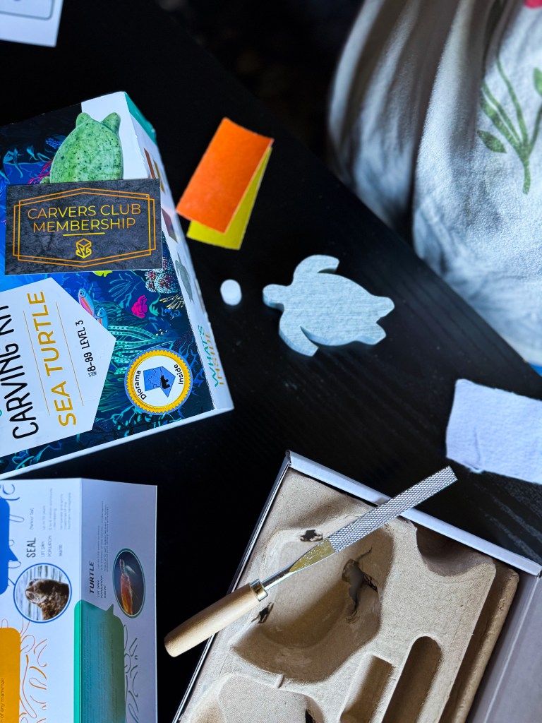 An assortment of carving materials laid out on a black table, including a Carvers Club Membership box, a turtle template, colored felt sheets, a carving tool, and a small white carving piece.