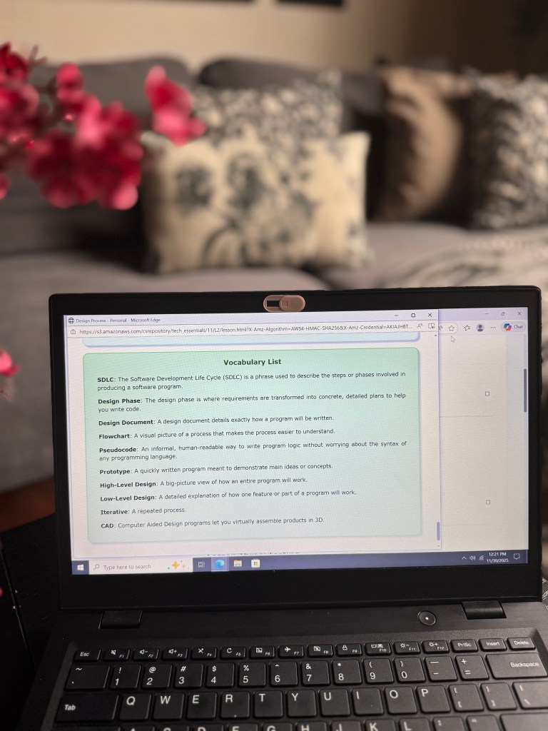 A laptop with an open web browser displaying a vocabulary list for a tech curriculum, including terms like SDLC, Design Phase, and CAD, with a soft background of a couch and floral decoration.