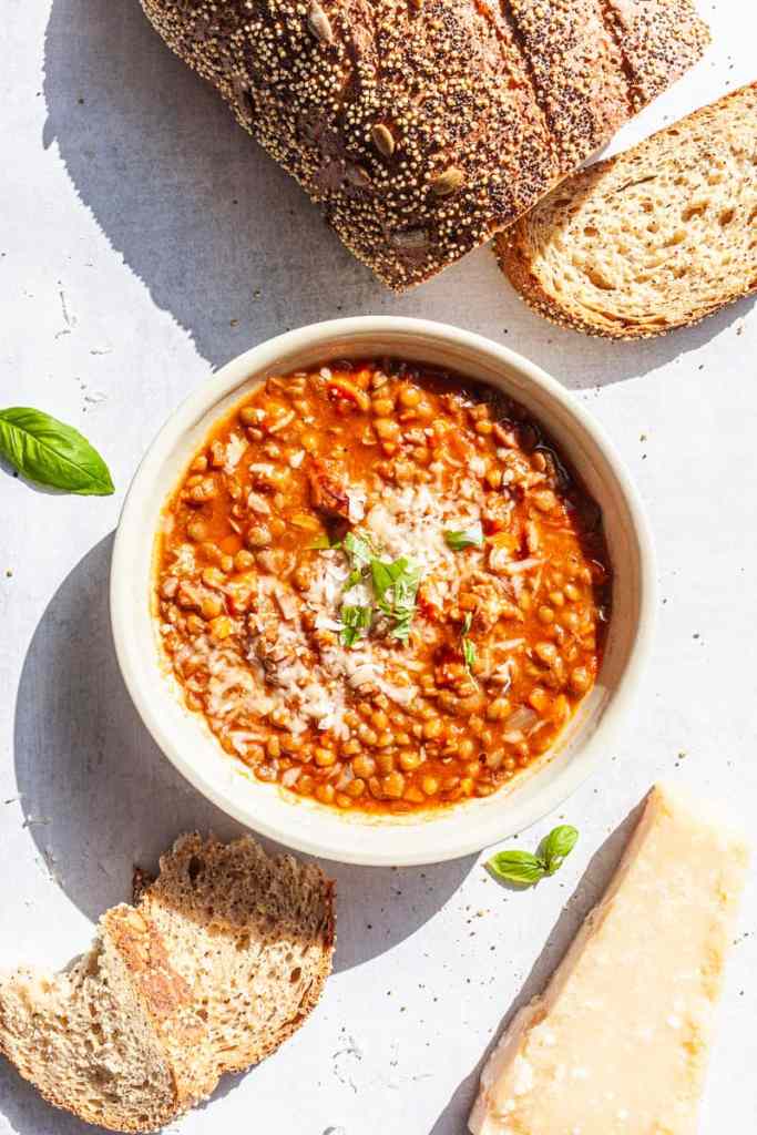 A bowl of hearty lentil soup topped with cheese and fresh herbs, accompanied by slices of whole grain bread and a piece of parmesan cheese.